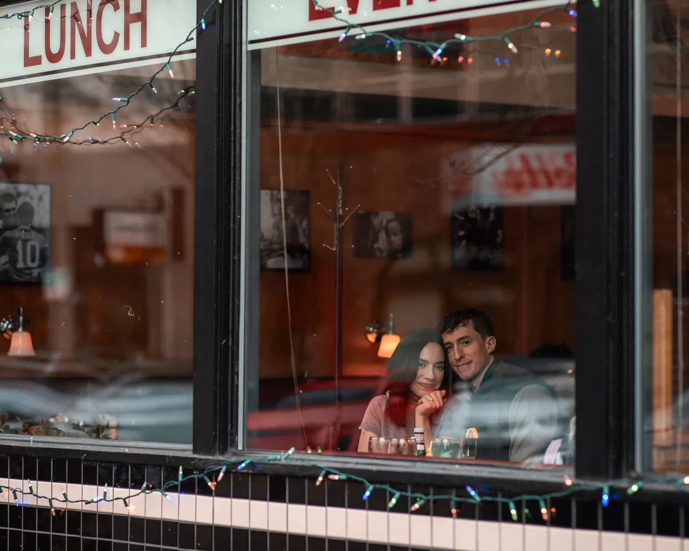 Documentary couple portrait seen through a restaurant window strung with holiday lights