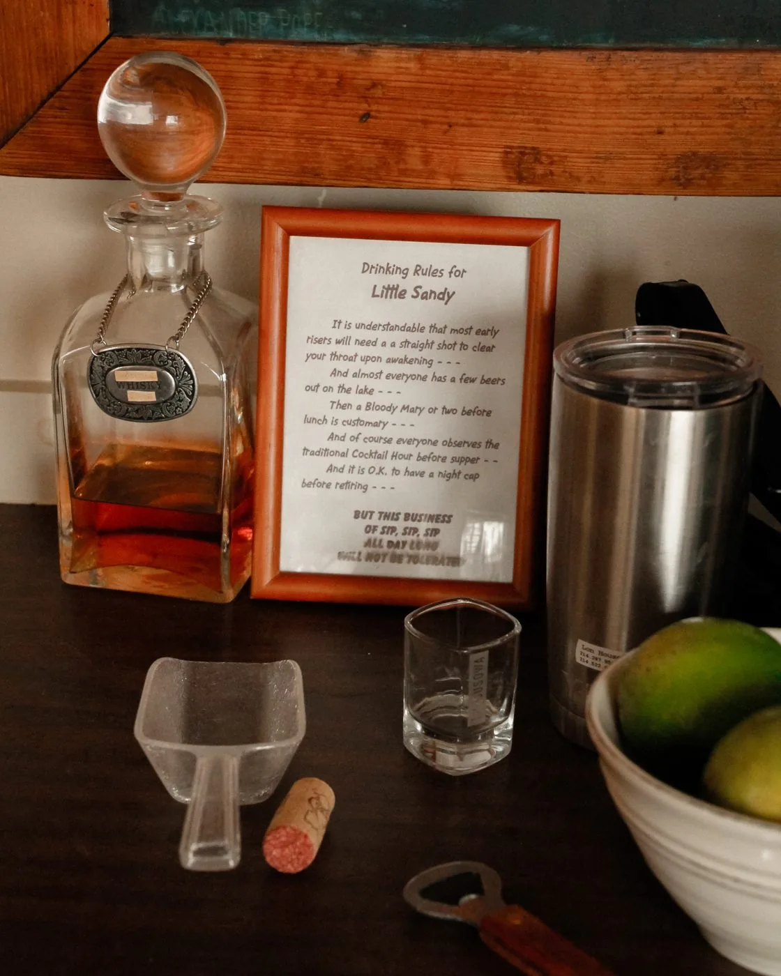 Documentary interior — cabin bar counter with framed drinking rules sign and glass decanter