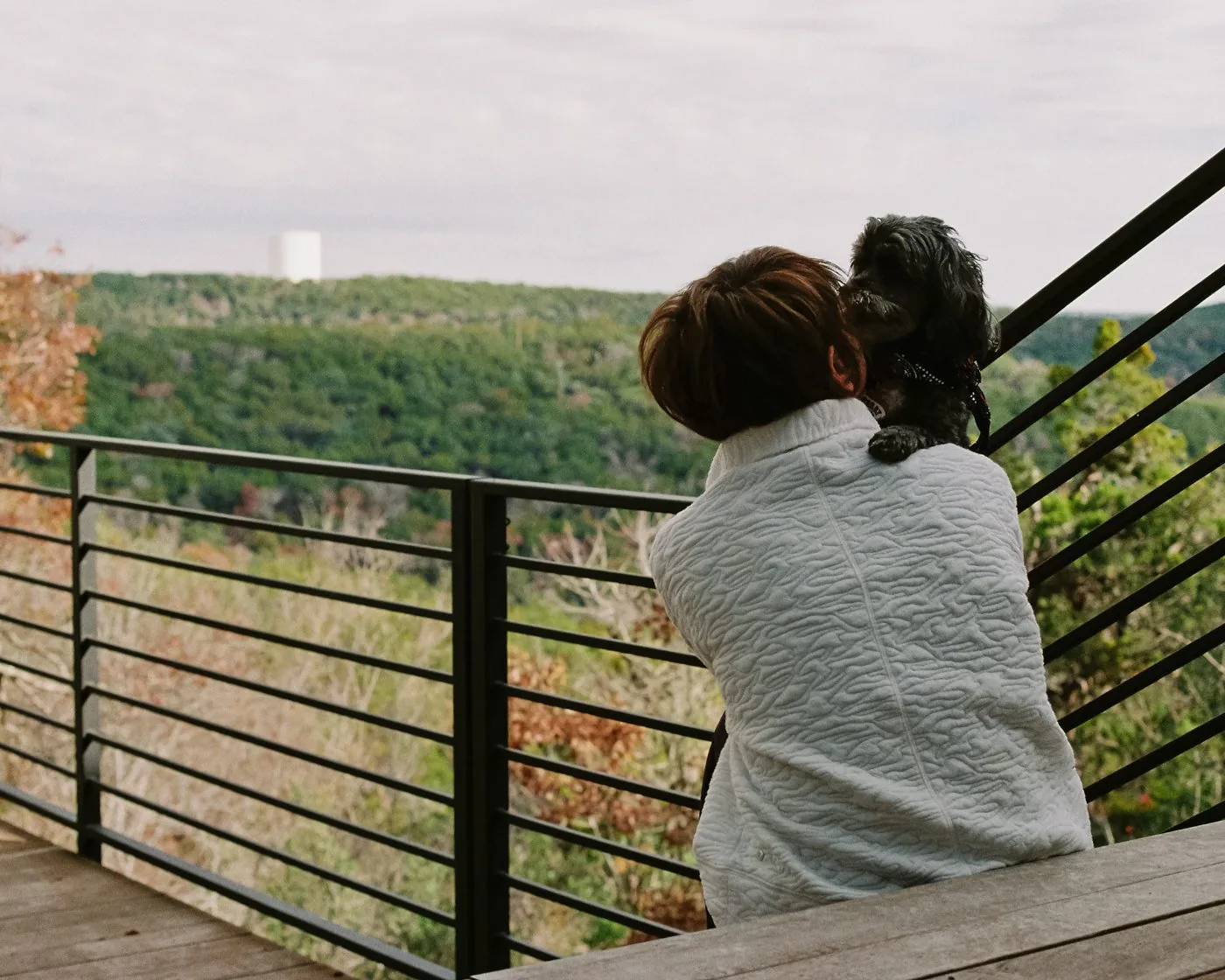 Documentary interior portrait — woman holding a small dog on a deck overlooking a wooded canyon