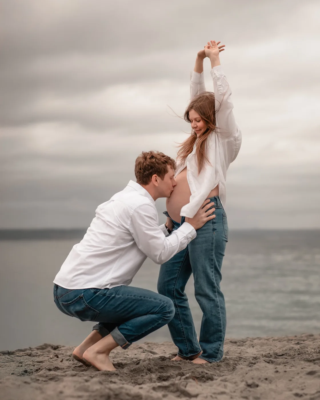Documentary maternity portrait — partner kneeling to kiss pregnant belly on the beach